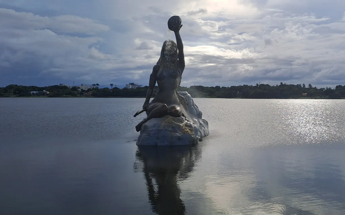 Estátua de uma mulher sobre uma pedra no meio de uma lagoa, refletida na água sob o céu nublado